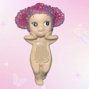 Puede incluir: Una figura caprichosa de una peque&ntilde;a figura de color nude con una cara sonriente y ojos grandes. La figura tiene una cabeza de material rosa y brillante. La figura se est&aacute; sosteniendo las manos cerca de la cara. El fondo es rosa claro con gr&aacute;ficos de mariposas y estrellas.