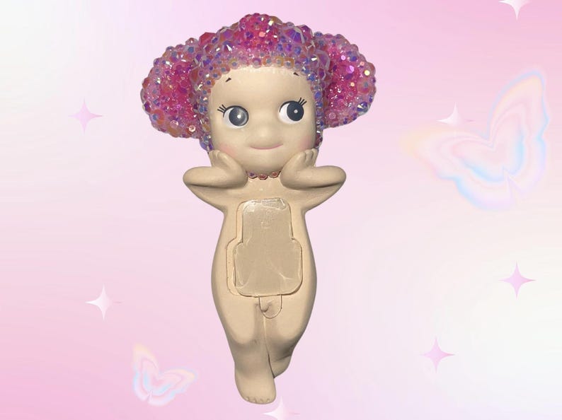 Puede incluir: Una figura caprichosa de una peque&ntilde;a figura de color nude con una cara sonriente y ojos grandes. La figura tiene una cabeza de material rosa y brillante. La figura se est&aacute; sosteniendo las manos cerca de la cara. El fondo es rosa claro con gr&aacute;ficos de mariposas y estrellas.