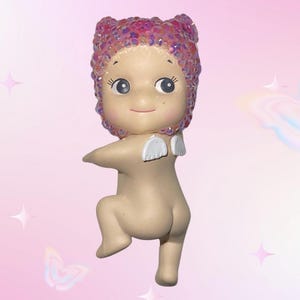 Puede incluir: Una peque&ntilde;a figura de beb&eacute; de color nude con una cubierta para la cabeza de lentejuelas rosas y peque&ntilde;as alas blancas. La figura est&aacute; en una postura juguetona, con una pierna doblada y los brazos extendidos. El fondo es un degradado rosa con estrellas y mariposas.