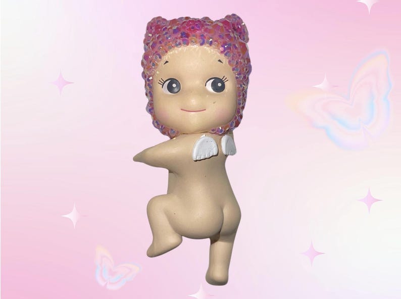 Puede incluir: Una peque&ntilde;a figura de beb&eacute; de color nude con una cubierta para la cabeza de lentejuelas rosas y peque&ntilde;as alas blancas. La figura est&aacute; en una postura juguetona, con una pierna doblada y los brazos extendidos. El fondo es un degradado rosa con estrellas y mariposas.