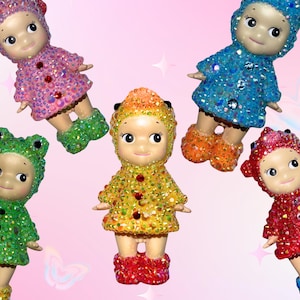Puede incluir: Seis peque&ntilde;as mu&ntilde;ecas decorativas en varios colores, incluyendo rosa, verde, amarillo, azul y rojo. Cada mu&ntilde;eca tiene adornos brillantes en su ropa y botas. El fondo es rosa suave con estrellas y mariposas.