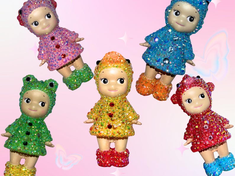Puede incluir: Seis peque&ntilde;as mu&ntilde;ecas decorativas en varios colores, incluyendo rosa, verde, amarillo, azul y rojo. Cada mu&ntilde;eca tiene adornos brillantes en su ropa y botas. El fondo es rosa suave con estrellas y mariposas.