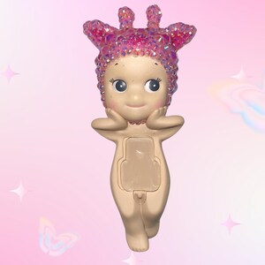 Puede incluir: Figura beige con un tocado rosa brillante y detalles a juego. La figura tiene una cara sonriente, los brazos levantados y un panel rectangular en la parte delantera. El fondo es un degradado de rosa con gr&aacute;ficos de estrellas y mariposas.