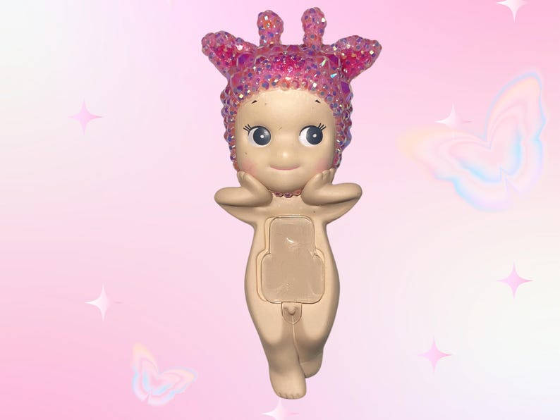 Puede incluir: Figura beige con un tocado rosa brillante y detalles a juego. La figura tiene una cara sonriente, los brazos levantados y un panel rectangular en la parte delantera. El fondo es un degradado de rosa con gr&aacute;ficos de estrellas y mariposas.