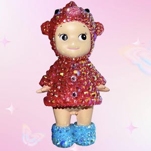 Puede incluir: Peque&ntilde;a figura de mu&ntilde;eca con un atuendo rojo enjoyado y una capucha a juego. La mu&ntilde;eca tiene botas azules enjoyadas y una cara sonriente. El fondo es rosa suave con acentos de estrellas y mariposas.