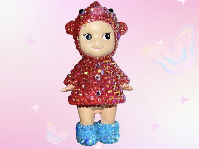 Puede incluir: Peque&ntilde;a figura de mu&ntilde;eca con un atuendo rojo enjoyado y una capucha a juego. La mu&ntilde;eca tiene botas azules enjoyadas y una cara sonriente. El fondo es rosa suave con acentos de estrellas y mariposas.