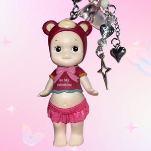 May include: A small doll wearing a red bear hat, a red shirt with a pink bow and the text "Be My Valentine", and a pink skirt. The doll is attached to a keychain with charms including hearts and a star.
