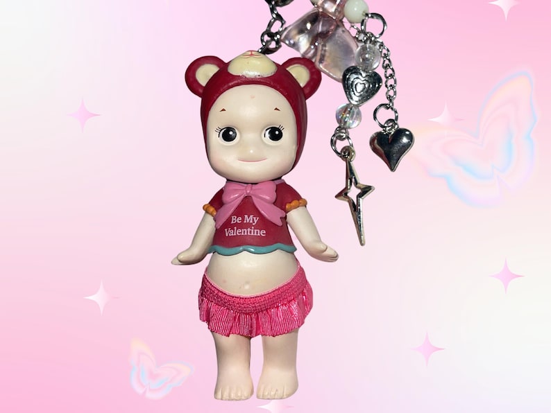 May include: A small doll wearing a red bear hat, a red shirt with a pink bow and the text "Be My Valentine", and a pink skirt. The doll is attached to a keychain with charms including hearts and a star.