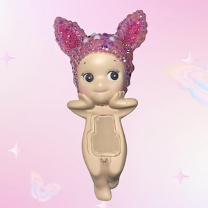 Puede incluir: Figura beige de un personaje querub&iacute;nico con ojos grandes y expresi&oacute;n serena. La figura lleva un tocado rosa con lentejuelas y orejas de conejo. El fondo es un degradado de rosa con acentos de estrellas y mariposas.