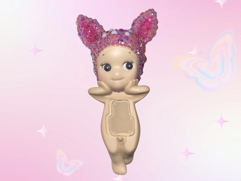 Puede incluir: Figura beige de un personaje querub&iacute;nico con ojos grandes y expresi&oacute;n serena. La figura lleva un tocado rosa con lentejuelas y orejas de conejo. El fondo es un degradado de rosa con acentos de estrellas y mariposas.