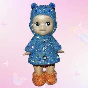 Puede incluir: Una peque&ntilde;a figura de mu&ntilde;eca con un abrigo azul con capucha y botas naranjas, ambas cubiertas de adornos brillantes. La mu&ntilde;eca tiene una expresi&oacute;n serena, sobre un fondo rosa.