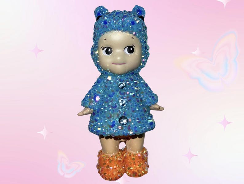 Puede incluir: Una peque&ntilde;a figura de mu&ntilde;eca con un abrigo azul con capucha y botas naranjas, ambas cubiertas de adornos brillantes. La mu&ntilde;eca tiene una expresi&oacute;n serena, sobre un fondo rosa.