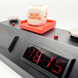 May include: A black digital timer with a red button and a white die with the words "Olde Towne" on it. The die is sitting on a red platform.