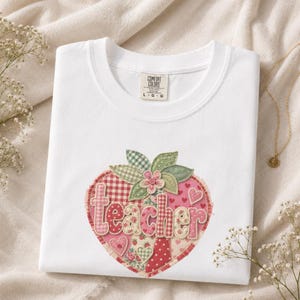 Könnte beinhalten: Ein weißes T-Shirt mit Rundhalsausschnitt und einem Comfort Colors Etikett in Größe L. Auf der Vorderseite befindet sich ein großes herzförmiges Motiv aus verschiedenen rosa und roten Stoffmustern. Das Wort teacher ist in hellrosa Buchstaben geschrieben.