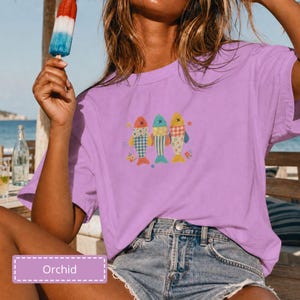 Puede incluir: Una camiseta de manga corta de color morado claro con un gráfico de tres peces estilizados con cuerpos estampados en rojo, azul y amarillo, acentuados por pequeñas estrellas y flores. La camiseta se combina con pantalones cortos de mezclilla azul.