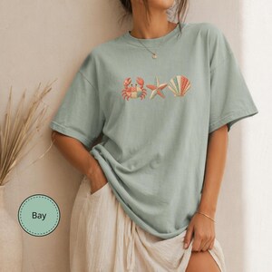 Puede incluir: Una camiseta de manga corta color verde salvia con un estampado gráfico de un cangrejo, una estrella de mar y una concha. El cangrejo, la estrella de mar y la concha tienen un patrón de patchwork multicolor. Una etiqueta circular muestra el texto Bay.
