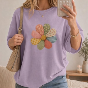 Op de afbeelding: Een lichtpaars t-shirt met een grote, veelkleurige bloemafbeelding op de borst. De bloem heeft bloemblaadjes met diverse patronen, waaronder bloemen, ruitjes en effen texturen in tinten roze, geel, groen en blauw, met een geel hart.