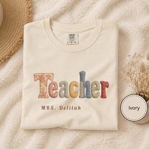 Puede incluir: Una camiseta de manga corta color marfil con la palabra Teacher en letras de tela multicolor con varios patrones florales y de cuadros. Debajo de la palabra, el texto MRS. Delilah está impreso en marrón. La camiseta tiene una etiqueta Comfort Colors.
