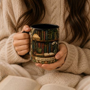 Puede incluir: Una taza de cerámica negra con una colorida ilustración de una estantería llena de libros, una lámpara encendida, una taza pequeña y un florero. El texto "Just one more chapter" está impreso en una banda decorativa en la parte inferior.