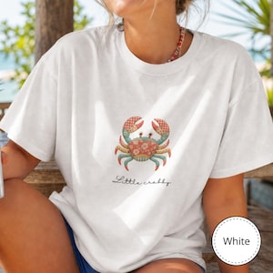 May include: A white short-sleeved t-shirt featuring a central graphic of a crab with a floral and checkered pattern in shades of red, blue, and cream. Below the crab, the text Little crabby is printed in a black, cursive, handwritten-style font.