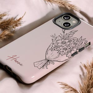 Puede incluir: Funda de teléfono con un fondo degradado rosa y un diseño de ramo floral negro. El nombre "Hanna" está escrito en cursiva. La funda tiene un protector de lente de cámara negro.