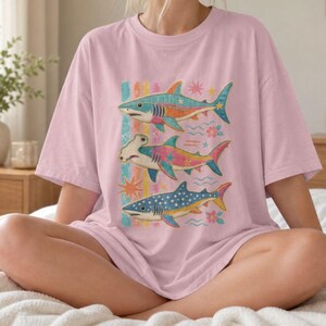 Op de afbeelding: Een lichtroze T-shirt met korte mouwen en een grafische print van drie verticaal gestapelde haaien. De haaien zijn afgebeeld in kleurrijke, patronen met sterren, bloemen en golvende lijnen op een lichtroze achtergrond.