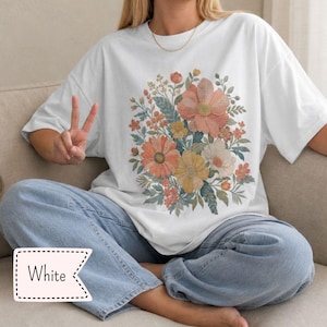 Op de afbeelding: Een wit T-shirt met korte mouwen en een centrale grafische print van een bloemenboeket. Het ontwerp bevat diverse bloemen in tinten perzik, oranje, geel en gedempt groen blad. Het shirt heeft een ronde hals en een losse pasvorm.