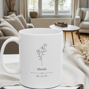 Puede incluir: Taza de cerámica blanca con un diseño floral negro, el nombre "Mariah" y la fecha "02-11-95" impresos. La taza tiene un asa curva. El fondo está borroso, sugiriendo un ambiente interior.