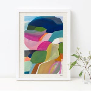 a vibrant abstract painting with a variety of colors and shapes, including shades of blue, pink, green, and orange. The painting is framed and displayed on a white surface, with a small vase of flowers to the right side.