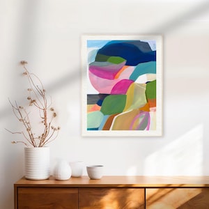 a vibrant abstract painting hanging on a white wall above a wooden dresser. The painting is a colorful abstract composition with various shapes and hues. On the dresser, there are three white vases and a small white bowl, adding a touch of elegance to the scene.