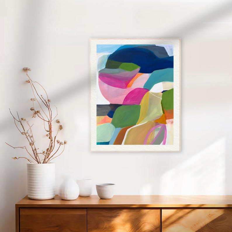 a vibrant abstract painting hanging on a white wall above a wooden dresser. The painting is a colorful abstract composition with various shapes and hues. On the dresser, there are three white vases and a small white bowl, adding a touch of elegance to the scene.