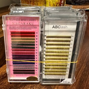 May include: Two clear plastic cases of false eyelashes. One case is labeled "NAGARAKU" with various colored lashes, including red, green, and blue. The other case is labeled "ABC lash" with green lashes. Both cases are secured with a yellow rubber band. The lashes are 11mm and 0.10C.