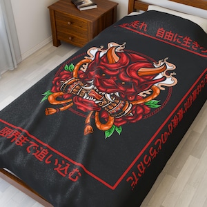 Könnte beinhalten: Schwarze Decke mit einem roten Oni-Masken-Design, Rosen und orangefarbenen Akzenten. Japanischer Text in Rot ist an den Rändern aufgedruckt. Die Oni-Maske hat detaillierte Merkmale und ist von einem kreisförmigen Design umgeben.