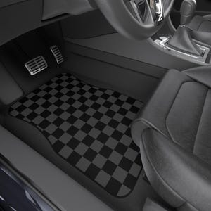 Op de afbeelding: Zwart en grijs geblokte automat. De mat is rechthoekig en past op de bestuurderskant van een voertuig. Het interieur van de auto is zwart, met een zwarte stoel, stuurwiel en versnellingspook zichtbaar.