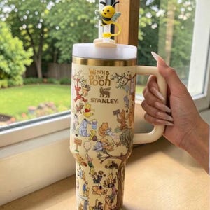 Pode incluir: Um copo Stanley bege com personagens de Winnie the Pooh. O copo tem uma tampa branca, uma alça creme e um adorno em forma de abelha amarela. O copo é decorado com ilustrações de Pooh e seus amigos.