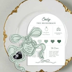 May include: A white plate with a gold rim holds a wedding-themed card and a ribbon-shaped tag. The card reads "Emily, The Bridesmaid" and includes details about the wedding. The tag says "I can't tie the knot without you."