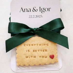 Puede incluir: Tarjetas de recuerdo de boda con los nombres "Ana & Igor" y la fecha "22.2.2025" impresos en negro. Una galleta con las palabras "EVERYTHING IS BETTER WITH YOU" y un corazón rojo está atada a la tarjeta con una cinta verde.