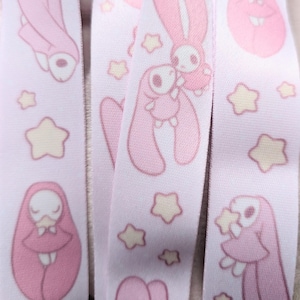 May include: Pink and white lanyard featuring a repeating pattern of cartoon bunnies, stars, and wrapped figures. The lanyard has a light pink background with the illustrations in shades of pink and cream.