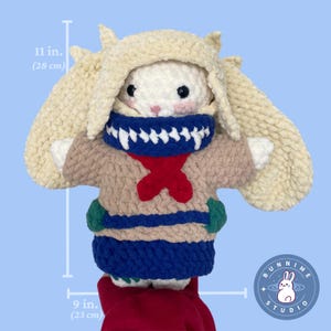 May include: A handmade, plush bunny doll, approximately 28 cm tall and 23 cm wide. The bunny wears a cream-coloured hood and a blue and red scarf. The doll has a tan sweater with green accents and a red bow.