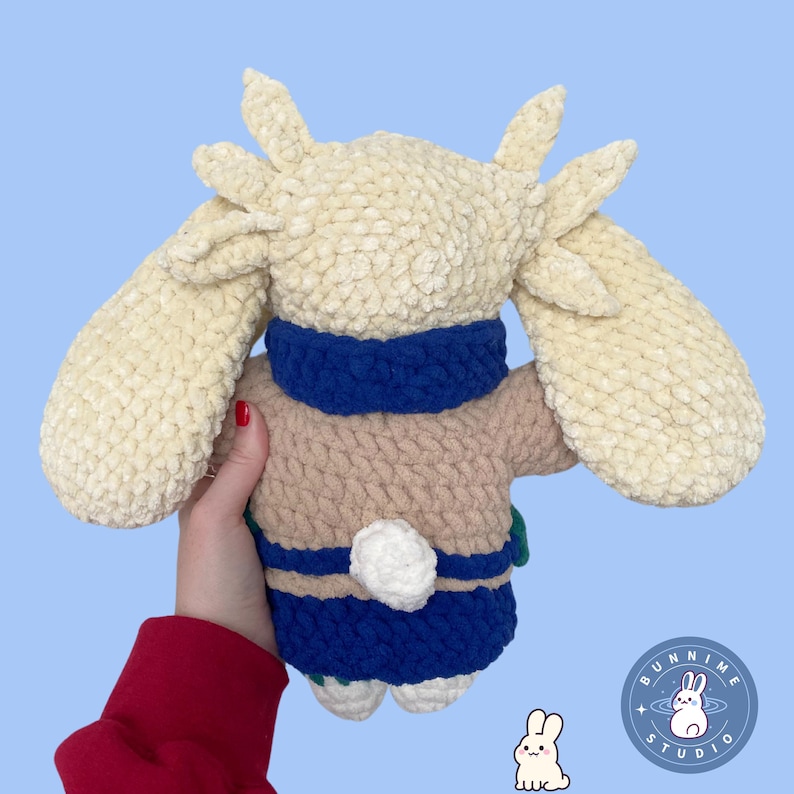 May include: A handmade plush bunny toy with long ears and a fluffy tail. The bunny is dressed in a blue and beige sweater with a blue collar. The toy is set against a light blue backdrop.