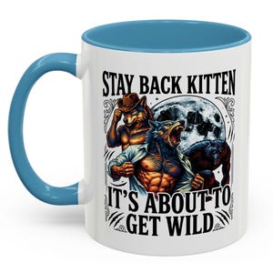 May include: A white ceramic mug with a light blue handle and rim, featuring a graphic of three anthropomorphic wolves in front of a full moon. The text reads STAY BACK KITTEN IT'S ABOUT TO GET WILD in black capital letters with decorative flourishes.