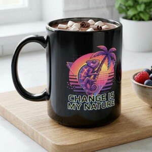 Op de afbeelding: Een zwarte keramische mok gevuld met een warme drank met marshmallows. De voorzijde toont een kleurrijke afbeelding van een kameleon op een palmboom tegen een zonsondergang met de tekst "CHANGE IS MY NATURE" in het wit gedrukt.