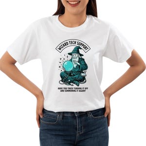 May include: A white t-shirt featuring a graphic of a wizard in a dark robe sitting cross-legged while holding a glowing blue crystal ball. The text reads WIZARD TECH SUPPORT and HAVE YOU TRIED TURNING IT OFF AND SUMMONING IT AGAIN with cables.