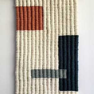 以下が含まれることがあります： クリーム色の背景に、錆、ネイビー、グレーの幾何学模様が施された織りの壁掛け。マクラメ作品は、吊り下げループと下部にフリンジが付いた木のダボに取り付けられています。