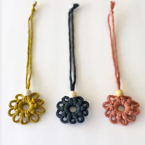 Può includere: Tre ornamenti floreali in macramè nei colori giallo senape, blu navy e rosa corallo. Ogni fiore ha un centro con perline di legno e un cordino abbinato per appenderlo. Gli ornamenti sono esposti su sfondo bianco.