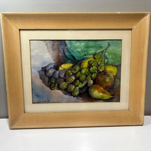 Könnte beinhalten: Aquarellmalerei eines Stilllebens mit Trauben, Birnen und Äpfeln. Das Obst ist in Grün-, Gelb- und Lilatönen dargestellt. Das Gemälde ist in einem hellbraunen Holzrahmen gerahmt. Die Signatur des Künstlers befindet sich unten links.