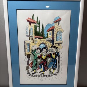 May include: A framed print depicting a festive scene with figures dancing in a town square. The artwork features a white frame, a blue mat, and a colorful illustration with buildings, trees, and people in traditional attire. The color palette includes red, green, yellow, and blue.