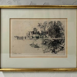 May include: A framed black and white etching on paper depicting a rural landscape with a pond, swimming ducks, grazing cattle, and distant buildings. The artwork is enclosed in a thin gold-toned metal frame with a wide off-white matting border.