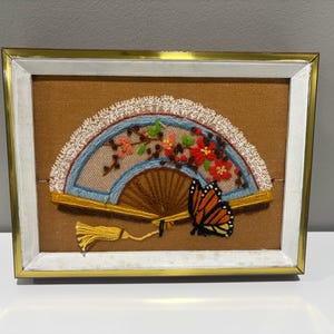 May include: Embroidered fan artwork in a gold and white framed display. The fan features a brown base with a blue and white lace trim, adorned with red flowers, green leaves, and an orange butterfly. A yellow tassel hangs from the fan.