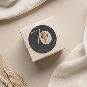 May include: A cream-colored gift box with a round sticker featuring a photo of a woman and the text "Michelle Johnson's 70th Birthday Party." The sticker is dark gray with gold accents. A cream ribbon wraps around the box.